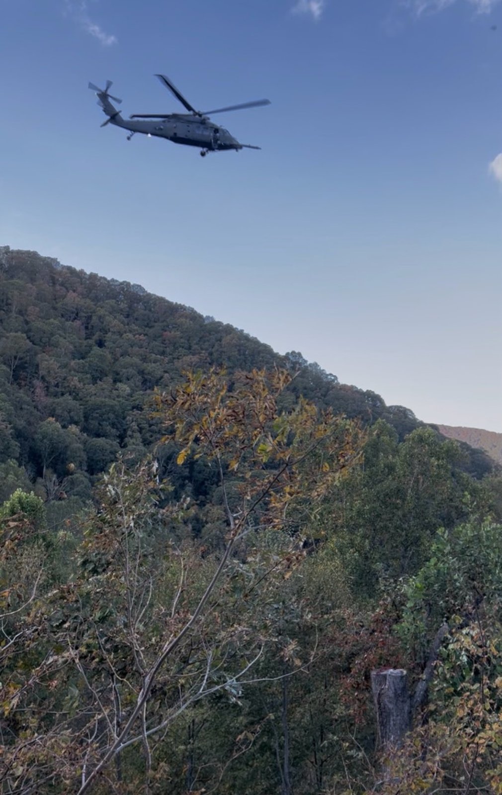 A military helicopter flies over a wooded ridge under a clear sky.