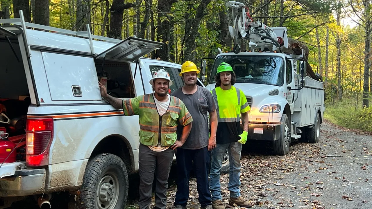 Three utility workers in hard hats stand beside a pickup and a bucket truck on a leaf-covered gravel road in the woods.