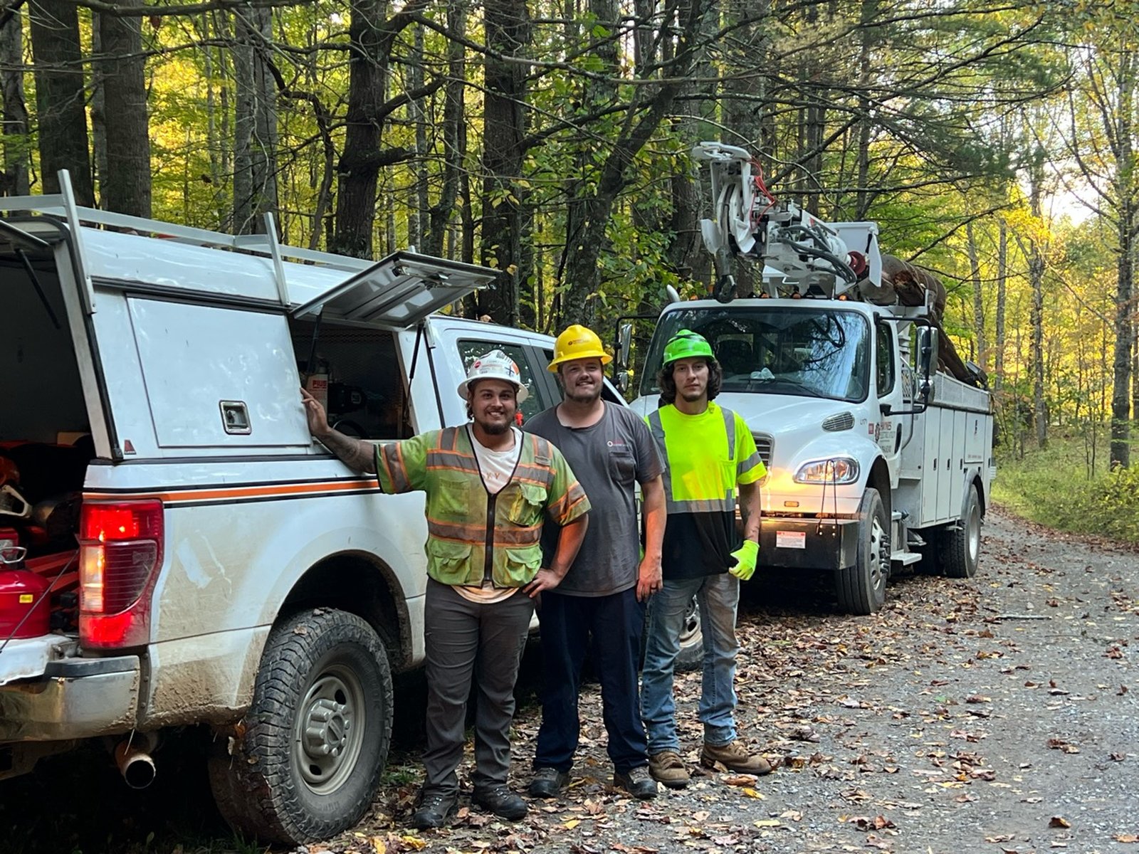 Three utility workers in hard hats stand beside a pickup and a bucket truck on a leaf-covered gravel road in the woods.